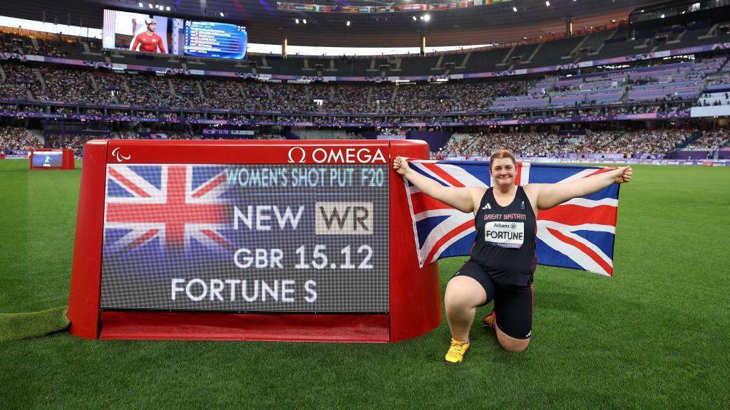 Sabrina Fortune holding flag in front of scoreboard showing her world record distance