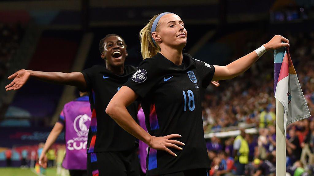 England striker Chloe Kelly stands calmly in front of the England fans after scoring the goal that sent them through to the final with one hand on top of the corner flag and her other on her hip, with team-mate Michelle Agyemang celebrating in the background