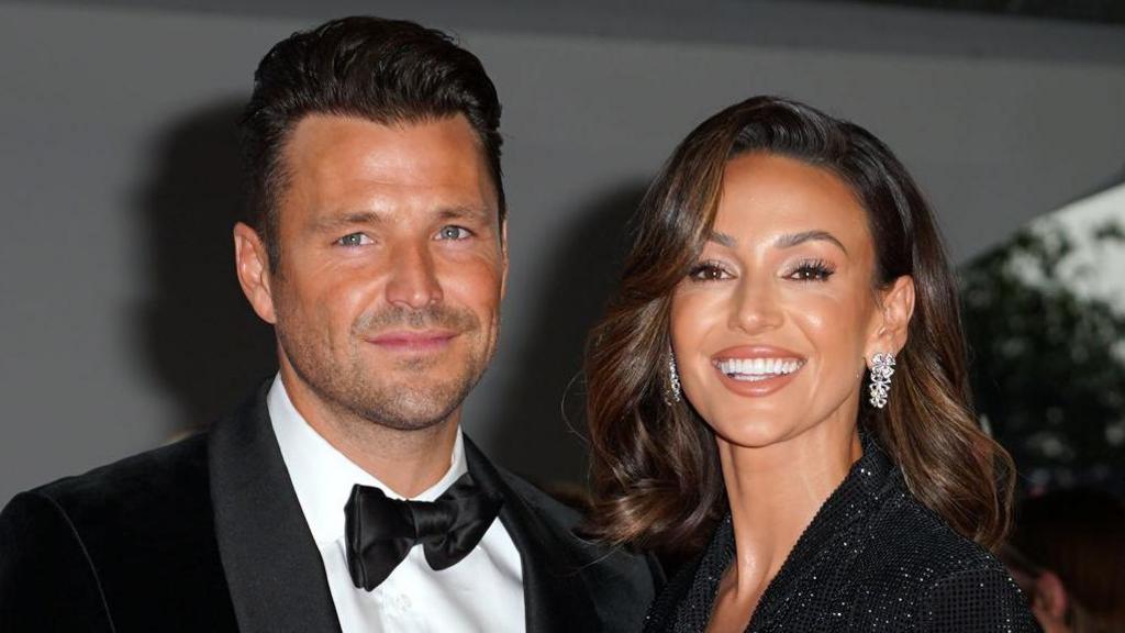 Mark Wright wearing a tuxedo while standing next to Michelle Keegan, who is wearing a sparkling black dress. Both are smiling.