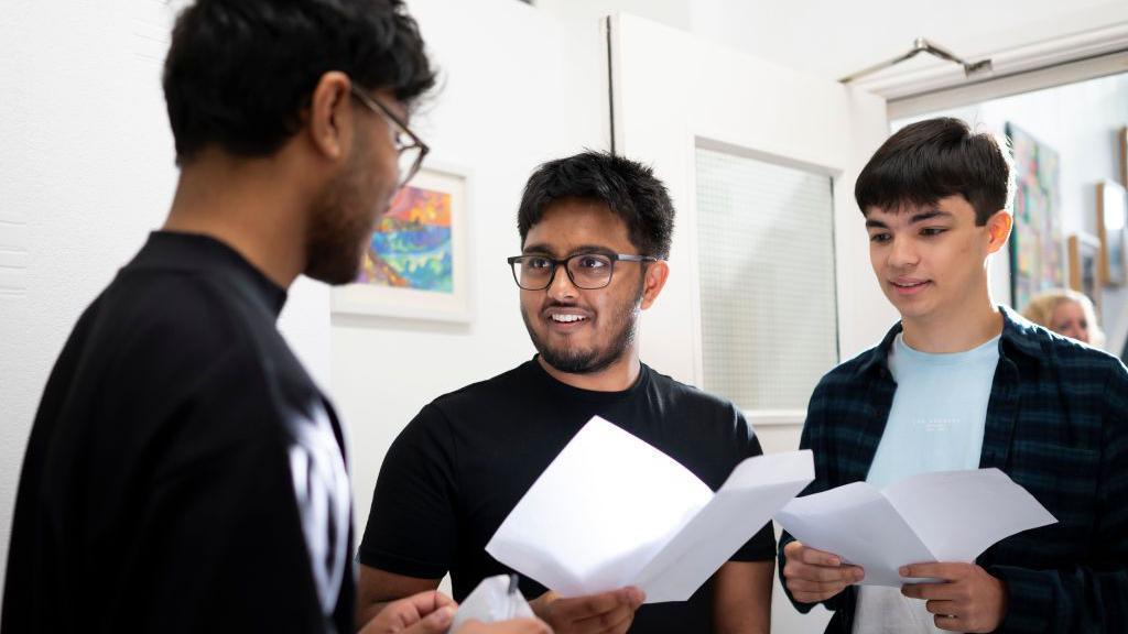 Habib Matlib, Umair Abdullah and John Bose receive their A-level results at Ffynone House School in Swansea, Wales. Umair is in the centre and is wearing a black T-shirt and black glasses - he is holding a sheet of paper with his exam results on and is looking at Habib, who is out of focus and can only be seen side-on. John is on the right of the three, wearing a pale blue T-shirt under a very dark blue checked shirt - both he and Umair are smiling.