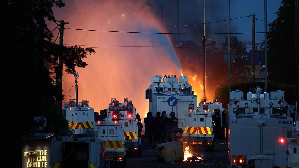 Police use water cannon as they block the road from crowds. A large number of police branded armoured vehicles block the road. There are pockets of fires seen between the vehicles and a stream from the water cannon.