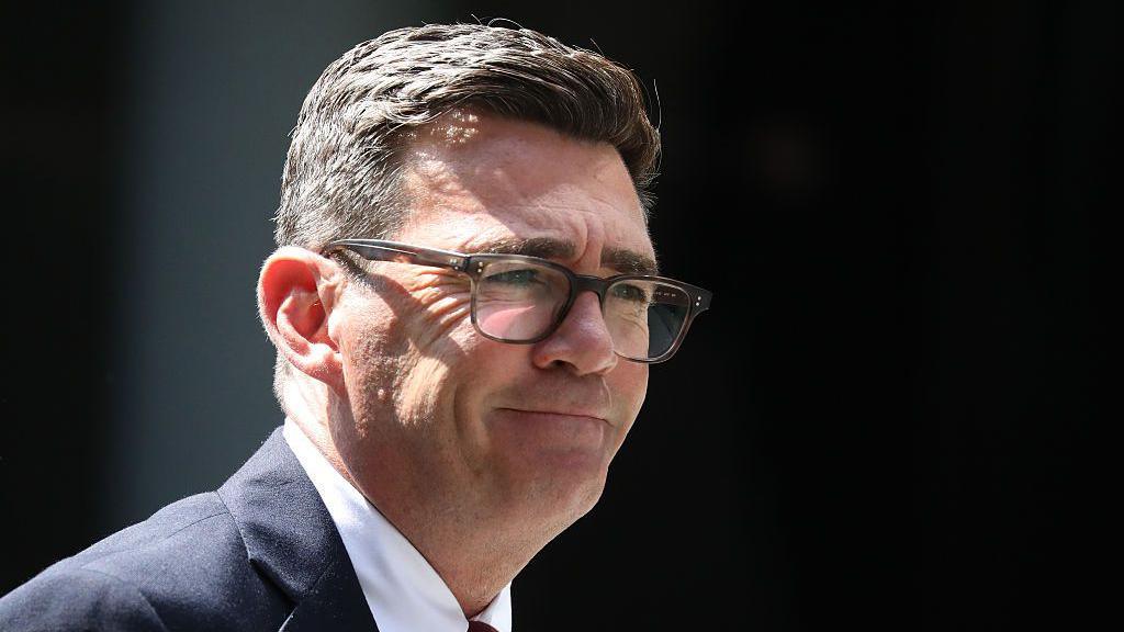 Andy Burnham is wearing glasses, a dark-coloured suit jacket, white shirt and a tie. He has a short, dark hair.