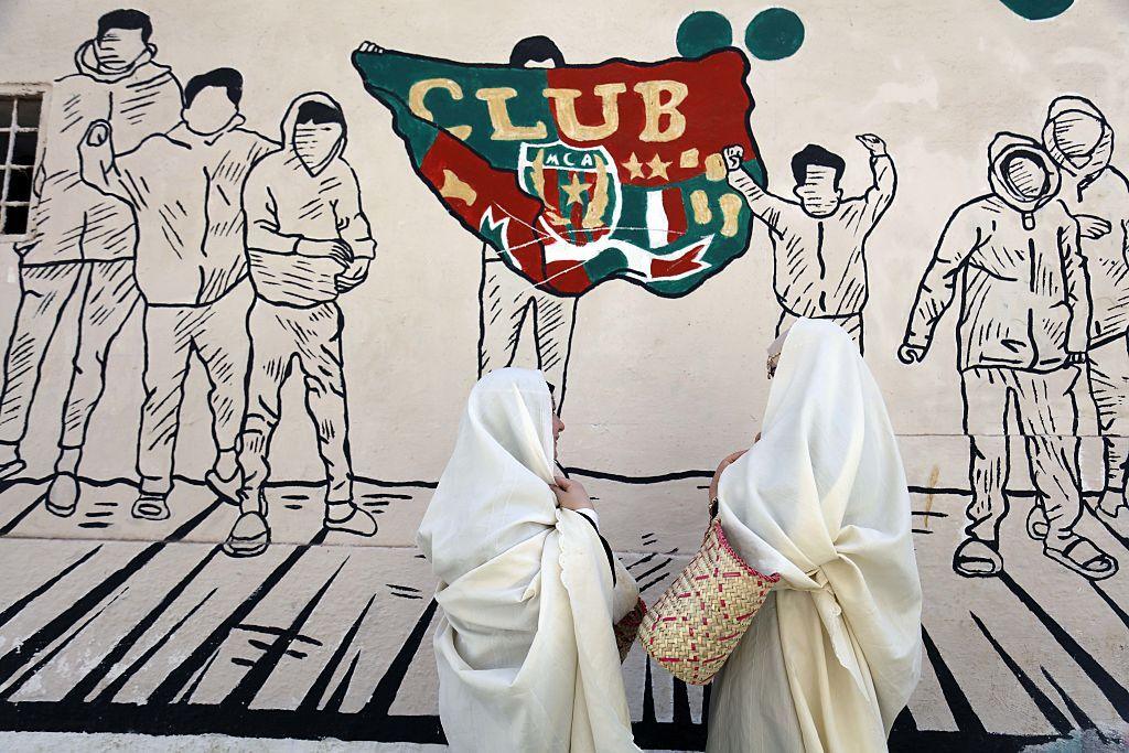 Two women stand in front of a mural wearing the haik - a modest gown covering most of the body typically worn in North Africa.