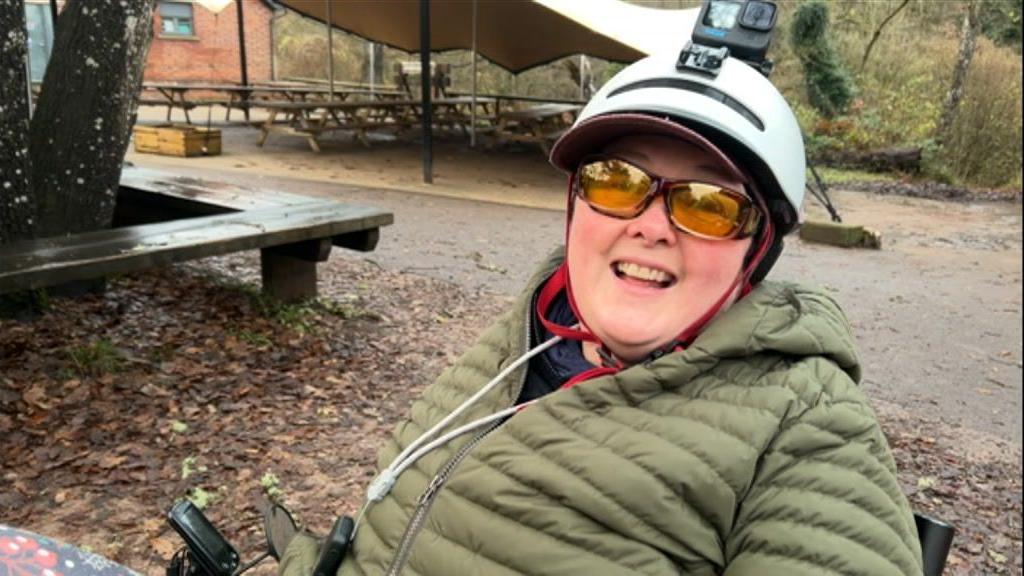 Debbie Cooper-Jones sitting in a recumbent bike in the Forest of Dean. It is an overcast day and there are brown leaves all over the ground. In the background there is an outdoor canopy with picnic benches underneath it. Ms Cooper-Jones is wearing a green padded coat, yellow sunglasses and a white helmet with a GoPro attached to the top. 