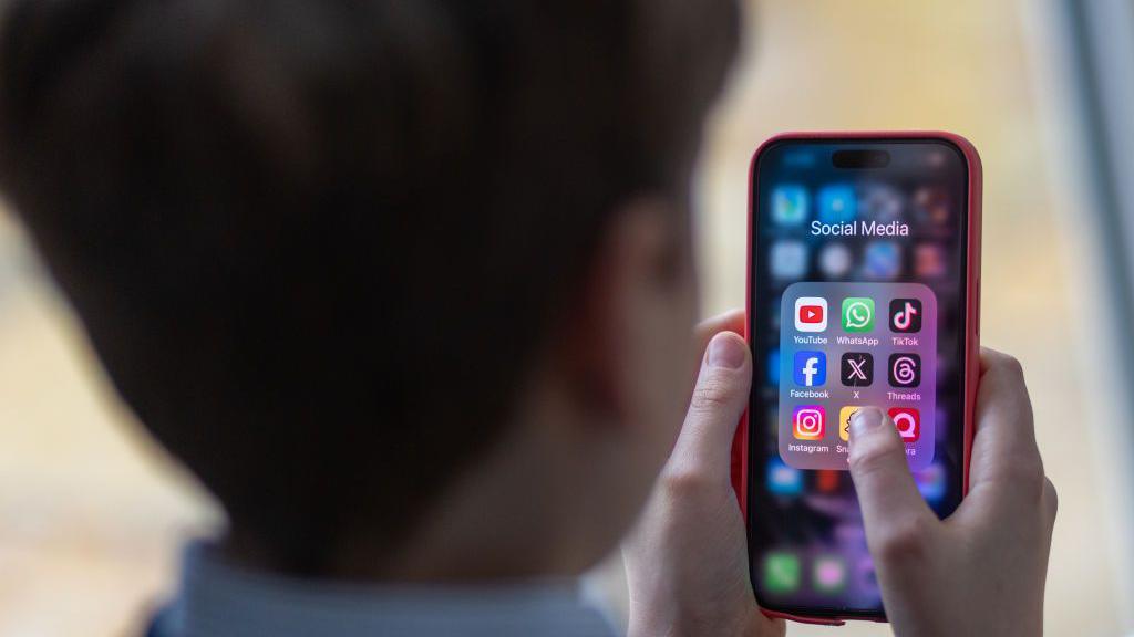 School boy looks at an iPhone screen showing various social media apps including TikTok, Facebook and X.