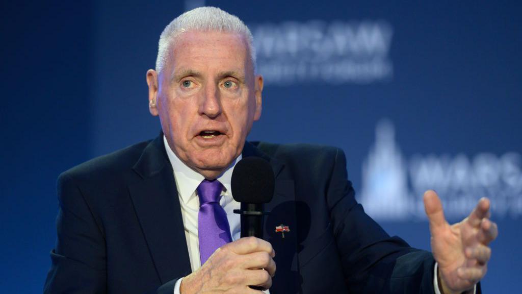 Lord Coaker speaking at an event. He is holding a microphone and has short grey hair. He is wearing a black suit with a white shirt and a purple tie. 