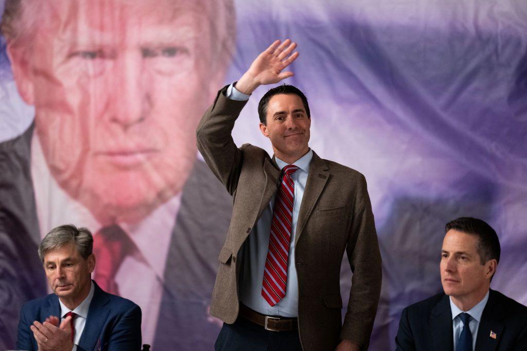 A man in a suit and tie standing and waving, flanked by two other men, the scene in front of a large picture of Donald Trump