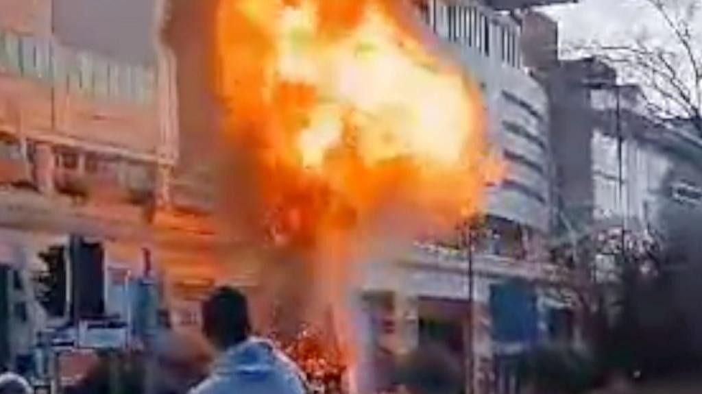 A phone image shows a large fireball errupting in front of a grey shopping centre with the back of an onlooker's head in the foreground 