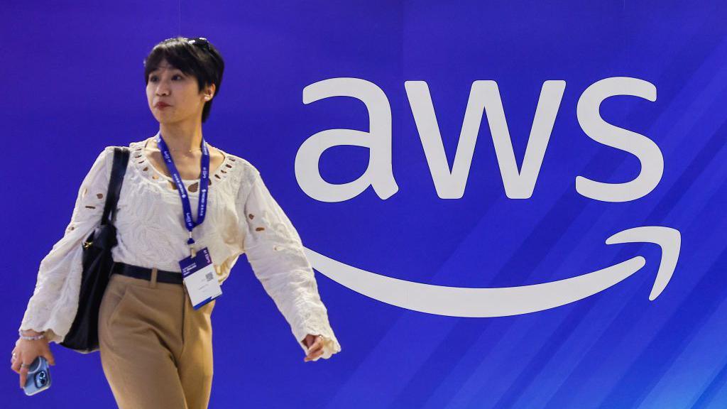 A person walking in front of an AWS sign. The sign is white text against a blue background. It is the letters AWS with a white arrow pointed in such a way it appears to make a smile.