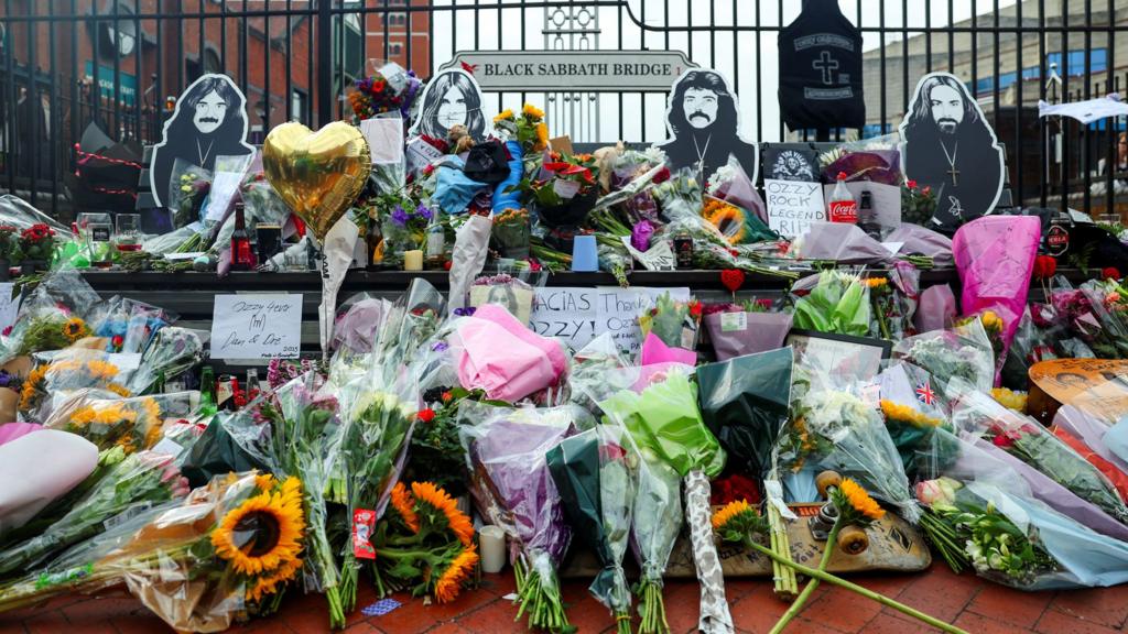 Flowers and other tributes bury the bench on Black Sabbath bridge in Birmingham, featuring likenesses of the four band members on the back of the bench