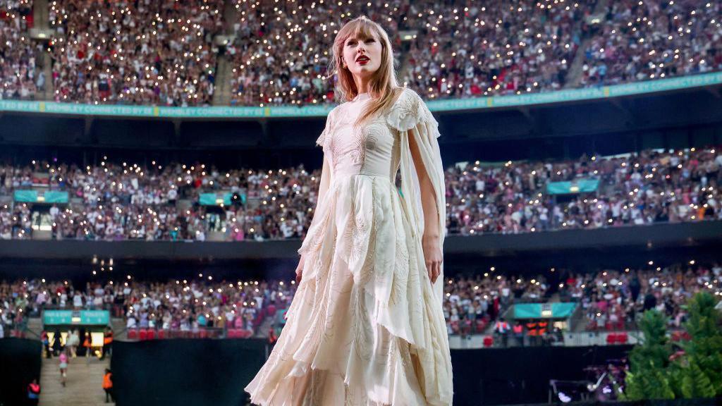 Taylor Swift in a white gown stands on stage with a large stadium crowd behind her.