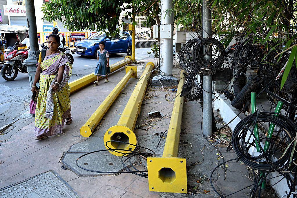 A woman walks past poles and wires lying on a pedestrian footpath in Chennai 