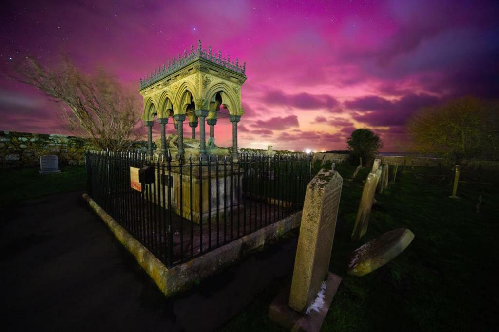 The purple and pink northern lights in the sky behind the Grace Darling monument which is a grey stone structure with decorative metal spike atop it.