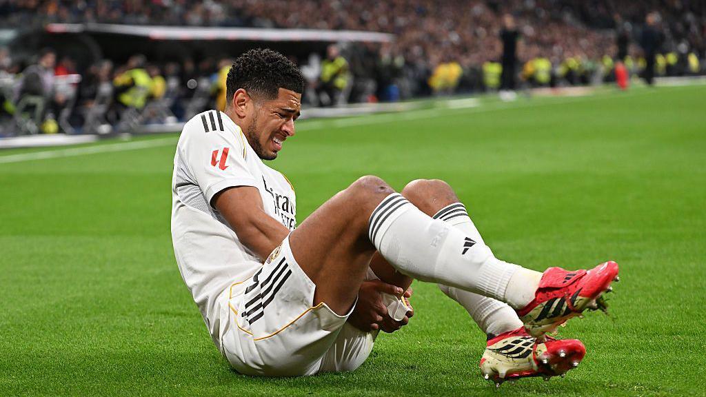 Jude Bellingham grimaces as he clutches his left hamstring with both hands. He has short black hair and is wearing Real Madrid's white home kit. His boots are pink.