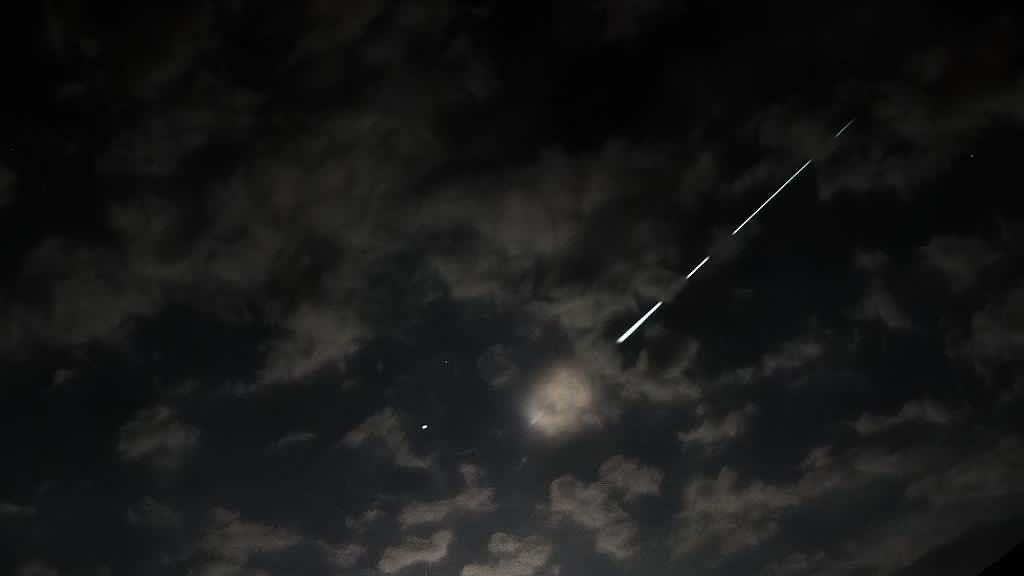 An Orionid meteor with a broken trail behind it in a dark, cloudy, moonlit night sky 