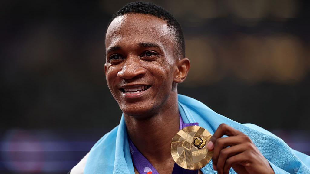 A headshot of Collen Kebinatshipi holding up a gold medal with his left hand as he poses for a photo while wearing the national flag of Botswana draped around his neck after winning the men's 400m final at the World Athletics Championships in Tokyo