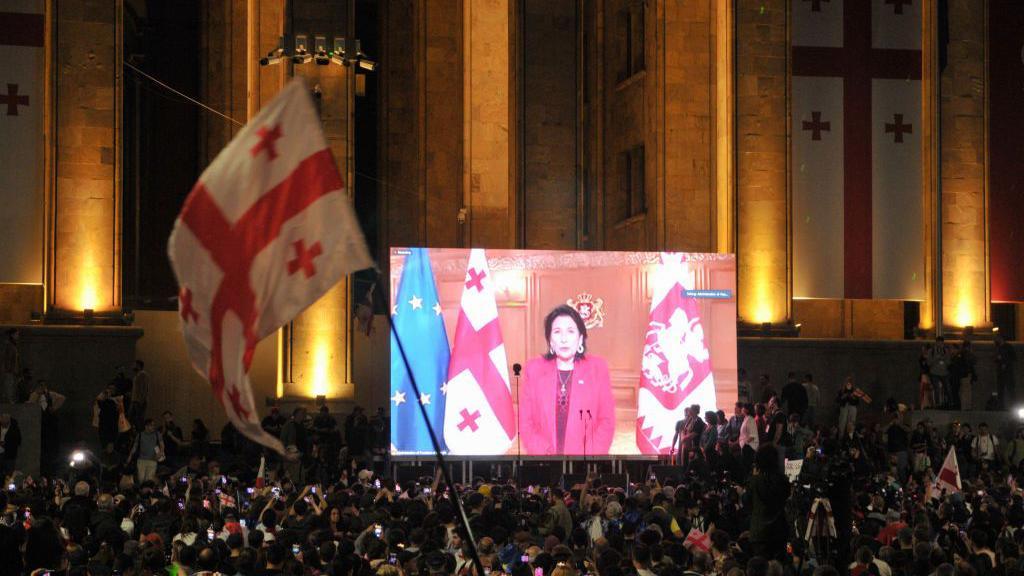 A large crowd gathers at night in front of the Georgian Parliament building in Tbilisi, with the illuminated facade adorned with Georgian flags. A giant screen displays President Salome Zourabichvili delivering a speech, standing between the Georgian and European Union flags.