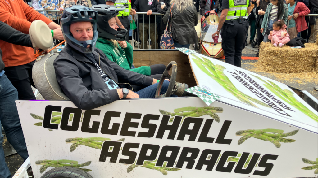 Two people are sat in a kart which is painted white and has asparagus on it. Across the side of the kart it says COGGESHALL ASPARAGUS in black writing. 