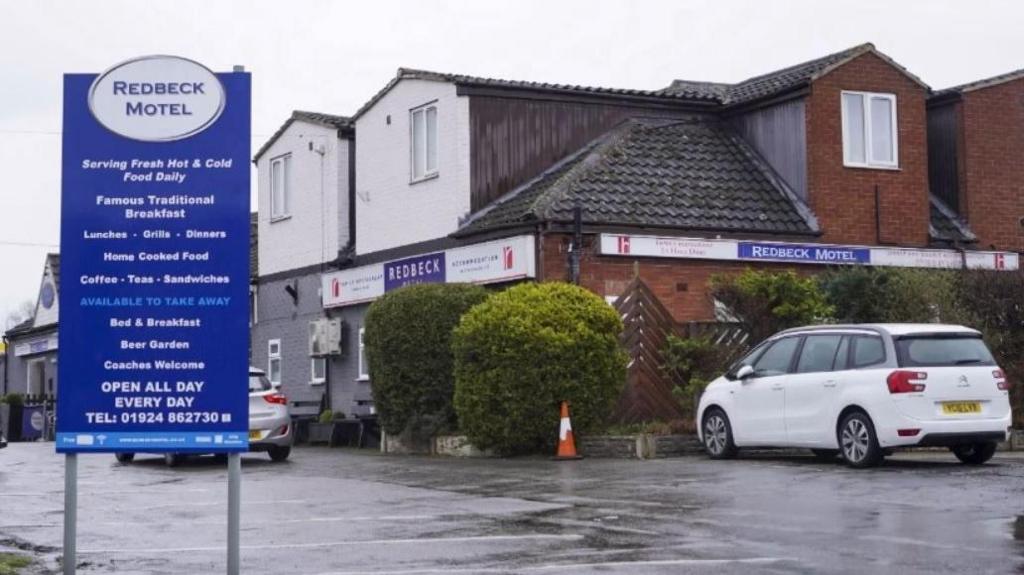 A motel building in a car park with a white and a silver car visible. There are bushes next to the building and a traffic cone, and a blue and white sign which reads 'Redback Motel'.