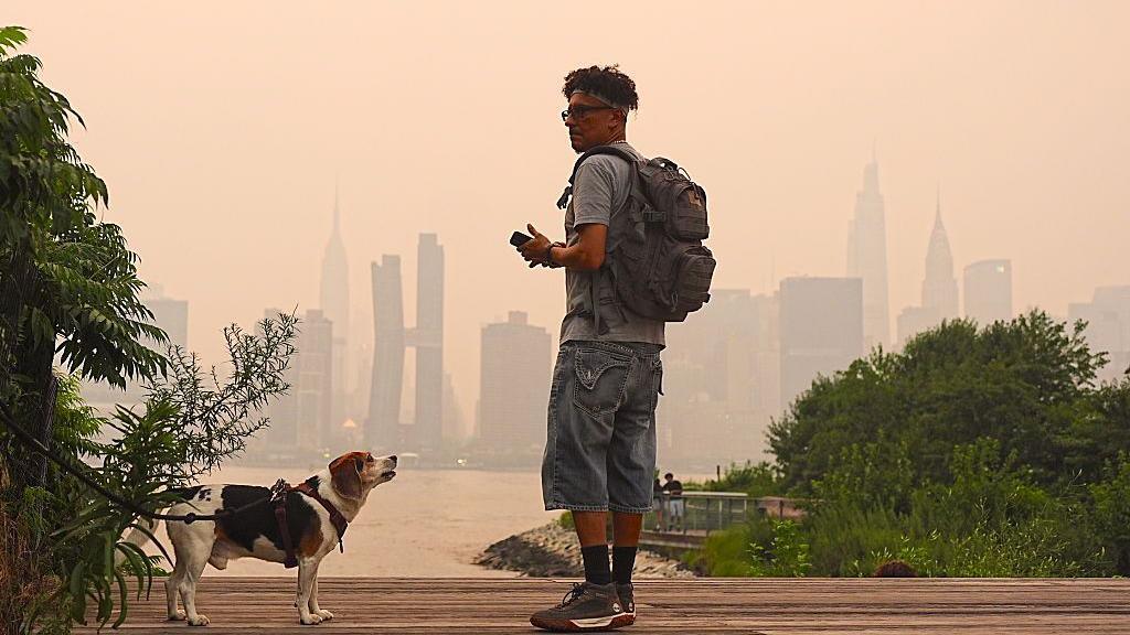 Man with backpack facing dog across river from hazy New York City skyline