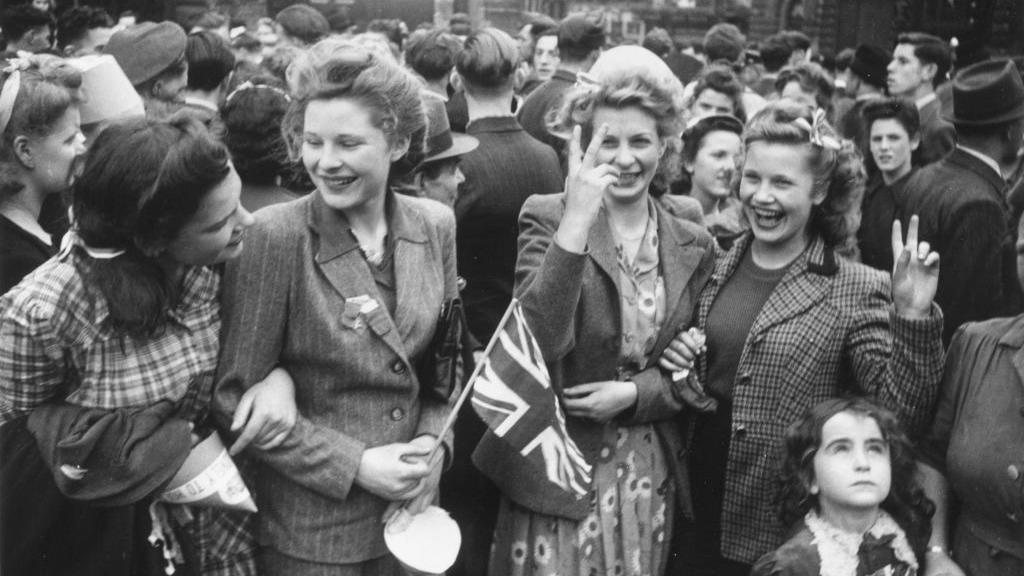 Four smiling women celebrating victory in 1945. They have linked arms and are smiling. One has a small Union Flag in her hand. A small girl is at the side looking up at the sky. 