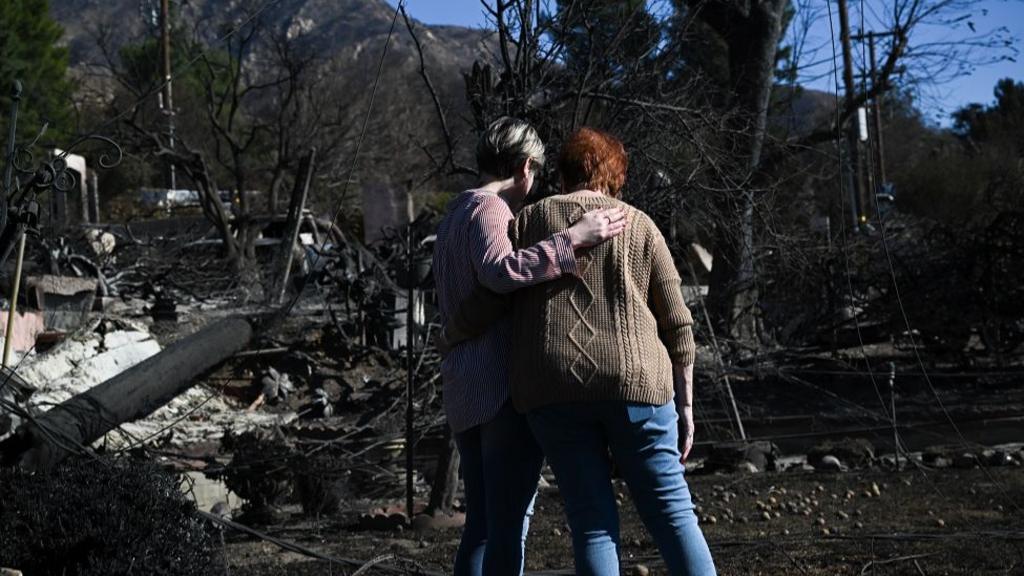 Two women stand and hold each other as they look at charred ground and the ruins of their home