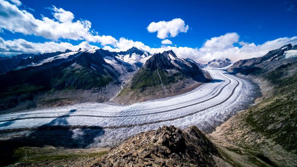 View of the Aletsch Glacier. Ice sweeps round from right to left, with mountains either side. Above is a blue sky with some clouds.