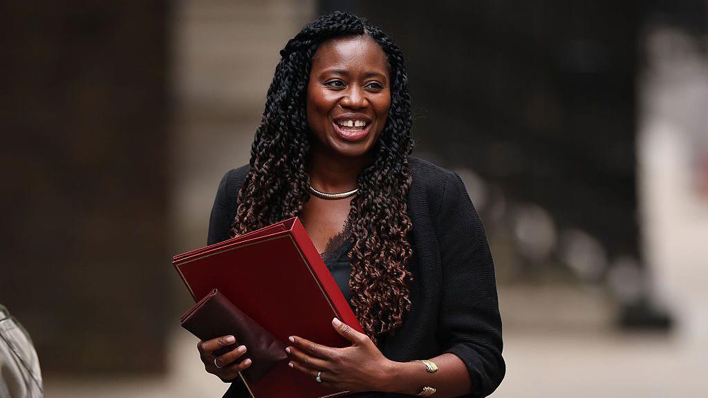 Miatta Fahnbulleh smiling and walking holding a red folder. She has long brown hair.