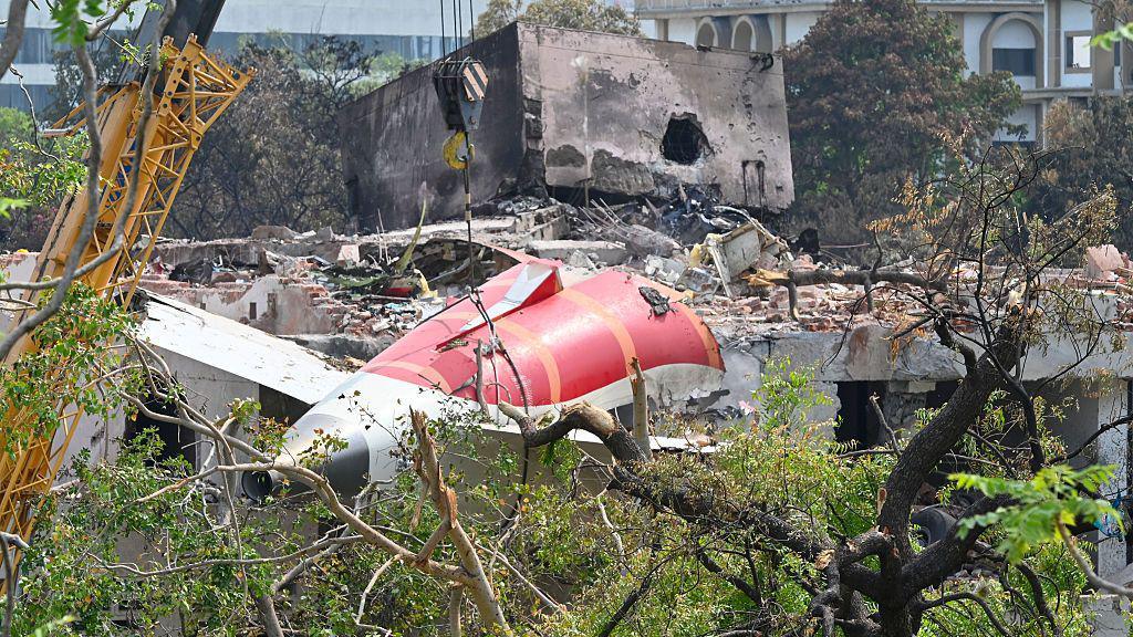 A crane lifts the tail of the wrecked Air India plane from the scene. Rubble and destroyed buildings are scattered around the ground.