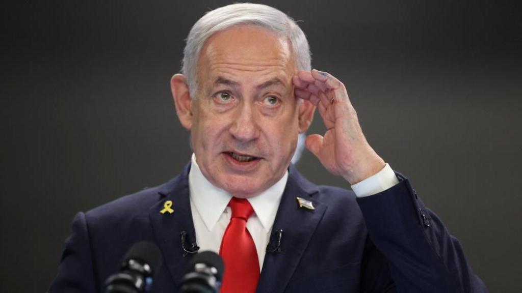 Netanyahu holding hand up with hand resting on side of his head, wearing blue suit and red tie.
