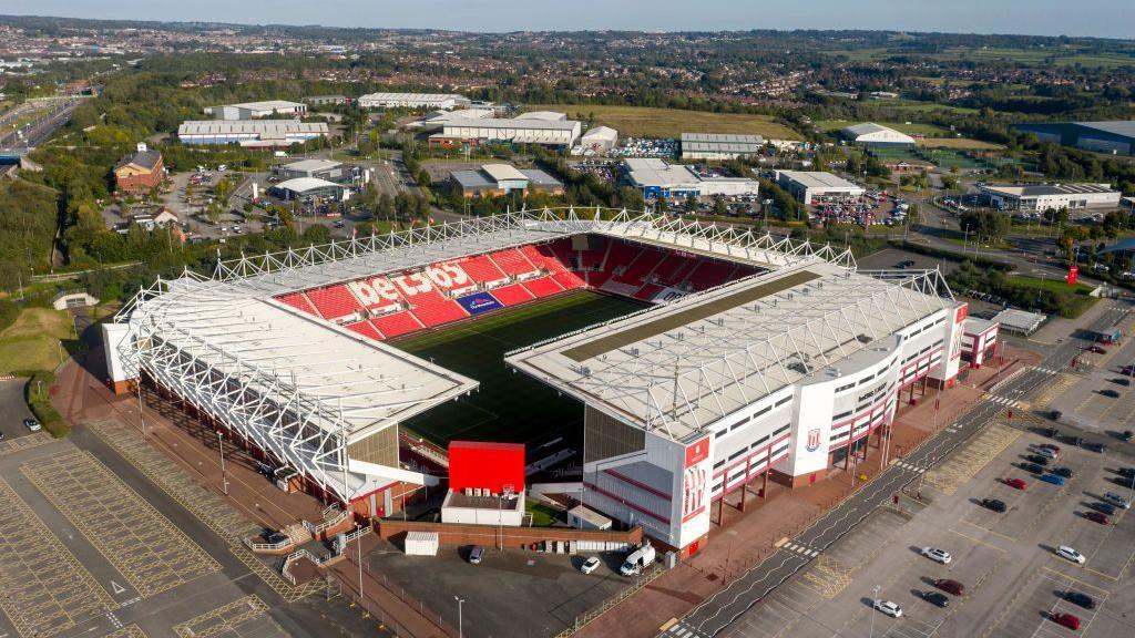 An aerial view of the Bet365 Stadium in Stoke-On-Trent. It has four stands and a car park. Rolling hills and homes can be seen in the background. 