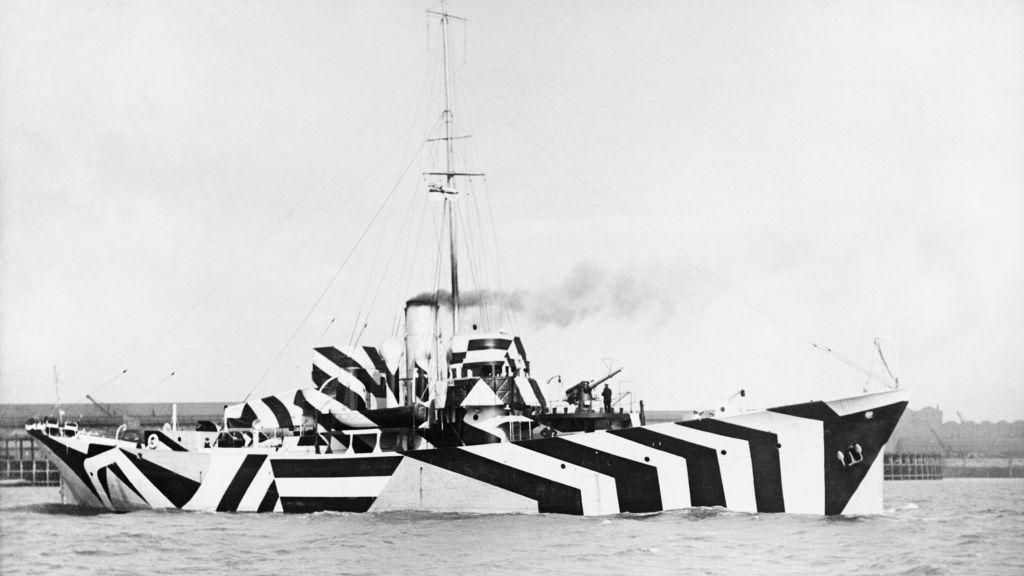 A black and white image of a large wartime ship in water with smoke coming out of a funnel. It is painted with a black and white geometric stripe pattern