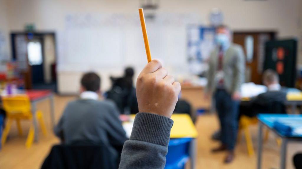 A classroom scence shows a child's hand raised, holding a yellow pencil. Other pupils and a teacher can be seen.