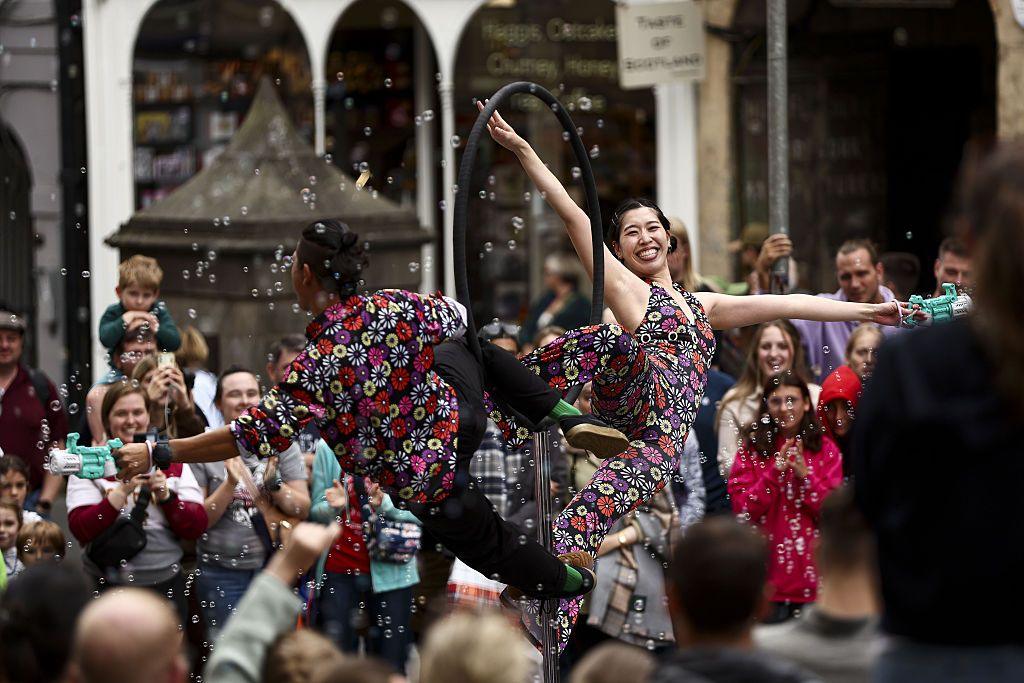 Male and female street performers in floral bright costumes pose on a metal ring prop with bubbles around them. People have gathered around them and are smiling and clapping.