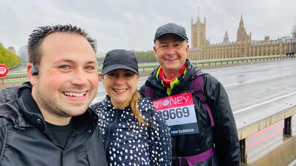Two men and one woman stood on London bridge in front of the Houses of Parliament. It's a grey day and one of the men is wearing a bib, which looks like he has taken part in a charity event, such as the London Marathon.