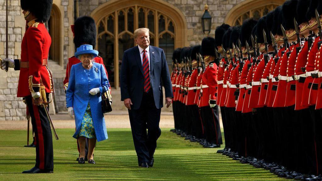 Donald Trump walks beside Queen Elizabeth II at Windsor Castle, he in a dark blue suit, and red-striped tie. She wears a bright blue suit-coat and matching hat. They are flanked by royal guards wearing the traditional red military coat, black pants and fluffy black hat.