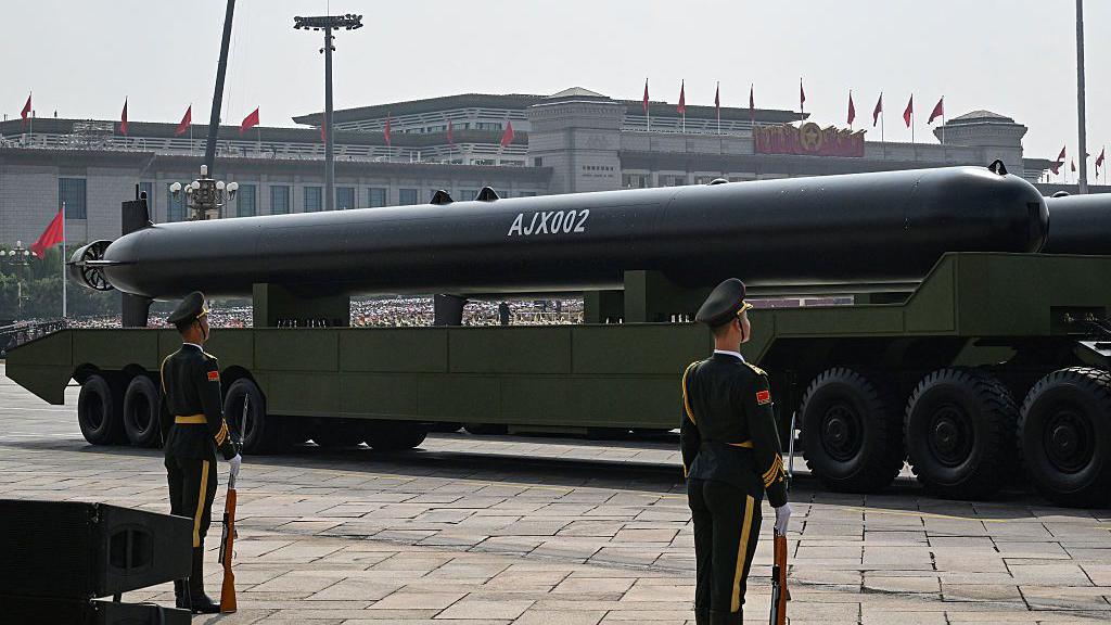 A large black missile on a flatbed truck, with AJX002 written in white paint on its side. Soldiers in ceremonial dress stand to attention in the foreground