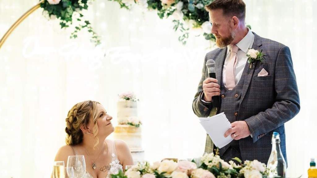 A groom looks at his new bride while making his wedding speech. He is wearing a grey suit and is holding a microphone in one hand and his notes in the other. A wedding cake stands in the background.