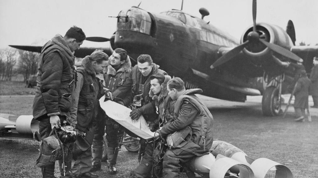 Six members of No 311 Squadron Royal Air Force Bomber Command - including a radio operator, a navigator, a pilot, a flight engineer and a front gunner - are studying a map on a bomb trolley which are about to be loaded  into their twin-engine Vickers Wellington MkIC medium bomber aircraft, which is behind them. The photo is in black and white.