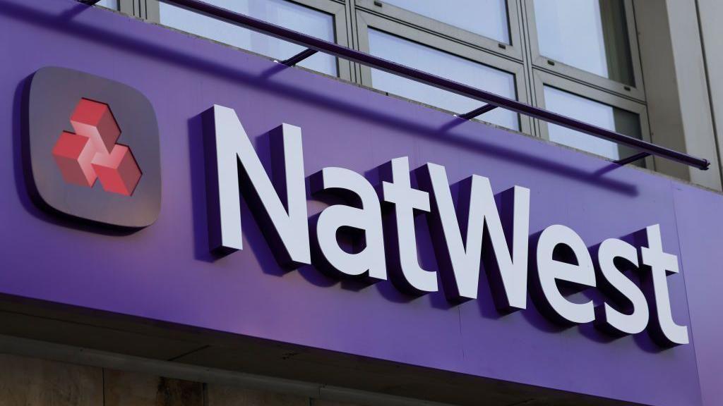 Close up of a NatWest sign. The background is purple and the text is white. It also features a small navy and red logo on the left.