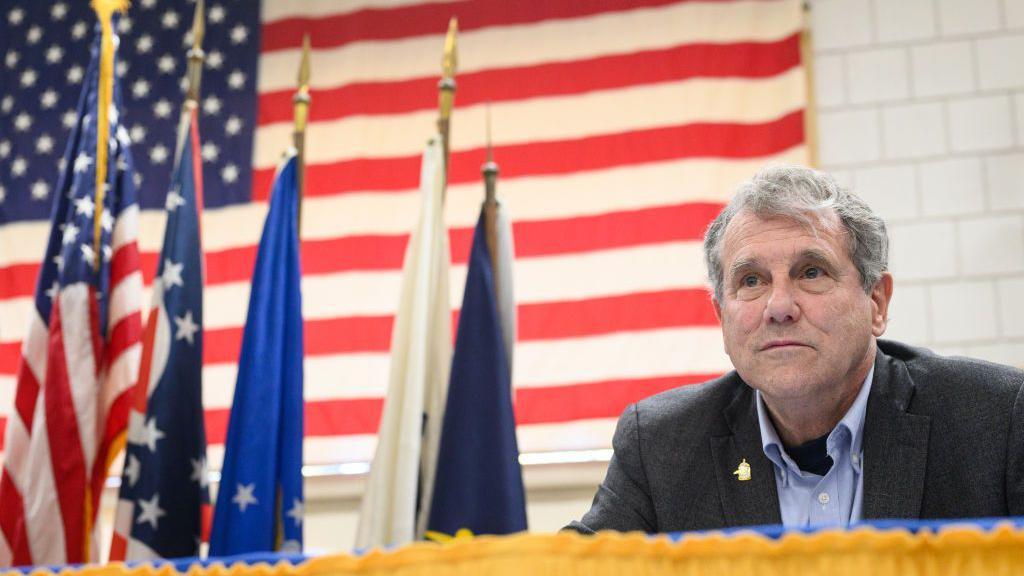 Sherrod Brown, wearing a black blazer with a golden pin on the right lapel, and a blue shirt, looks out into an unseen audience as he sits at a United Autoworkers event. Five flags on poles stand to his right with an American flag on the wall behind them.