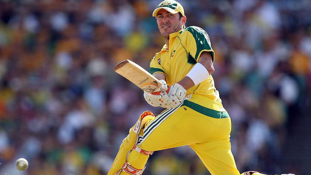 A man, wearing a yellow and green cricket uniform with yellow batting pads, hits a cricket ball with a bat. The ball is seen in the lower left corner.