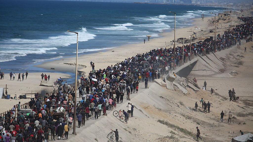Hundreds of Palestinians walk along a narrow coastal road to the north of the Gaza Strip. Most are walking on foot with their belongings. To their left is the beach and the sea. Some others are walking off road on a slope to the road's right.