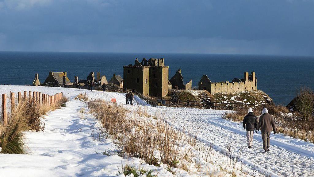 snow on the ground with people walking along a path close to a large castle like building next to a coast