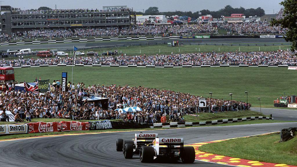 A rear view of the Williams of Nelson Piquet following closely behind team-mate Nigel Mansell during the 1986 race at Brands Hatch. Ahead of them, crowds can be seen snaking across the infield with a grandstand in the background