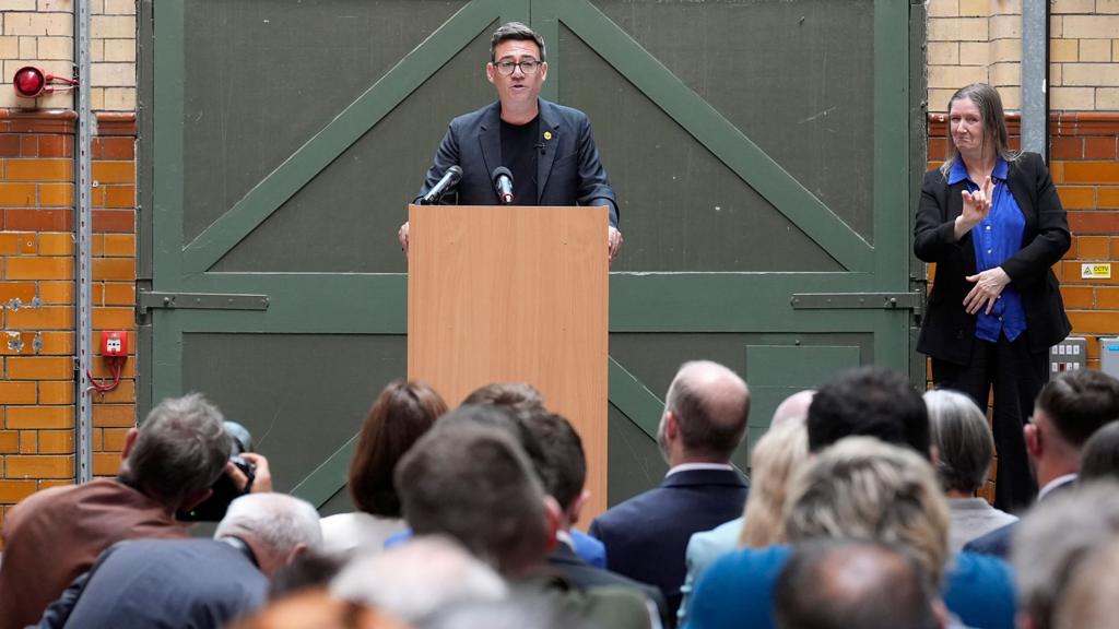 Burnham speaks at a podium with a sign language interpreter to his side and an audience in the foreground.