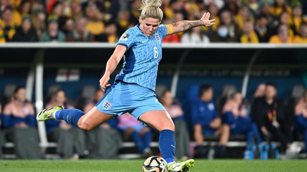 A woman in a bright-blue England football kit lunges to kick a football on a pitch.