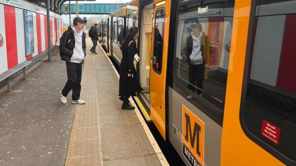 Two men and a woman board a black and yellow Metro train towards Newcastle Airport from Platform 2 at the Stadium of Light station in Sunderland.

