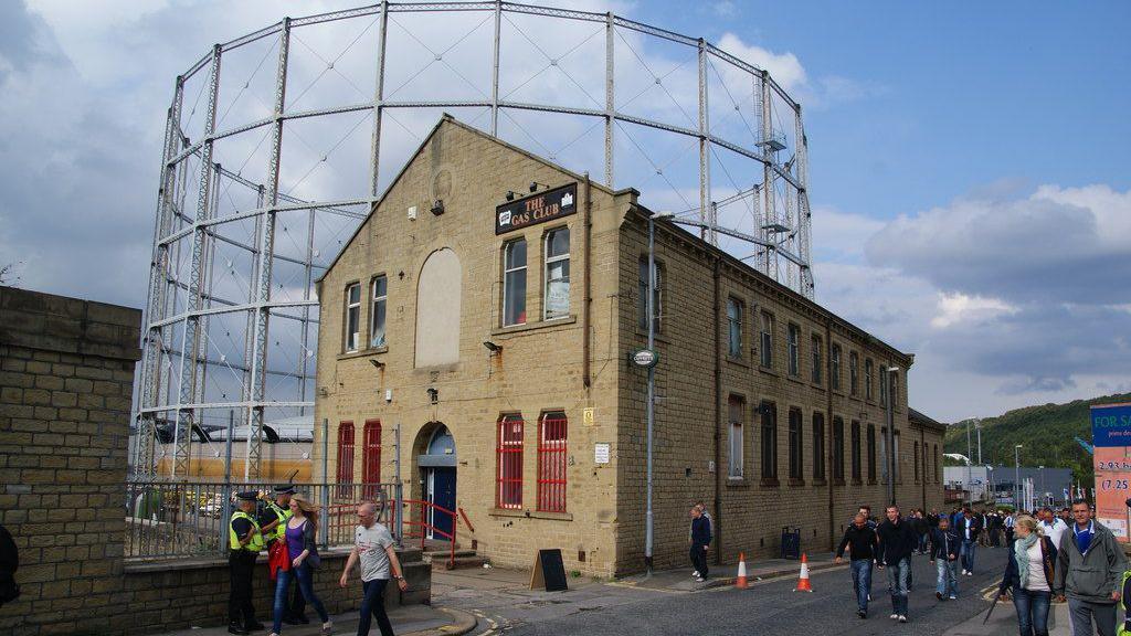 A two-storey venue in Huddersfield called The Gas Club, with football supporters walking past on the way to watch Huddersfield Town. In the background is a gas holder. 