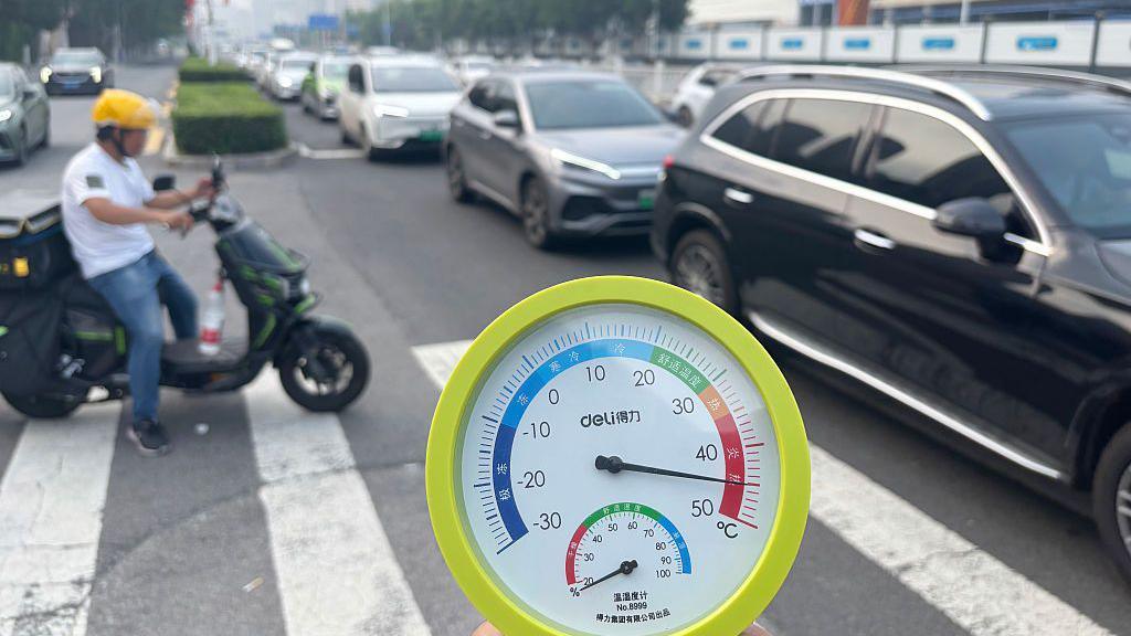 Close-up of a thermometer showing the temperature of the air exceeding 40 degrees Celsius. Behind it is a busy road with cars and a motorcycle.